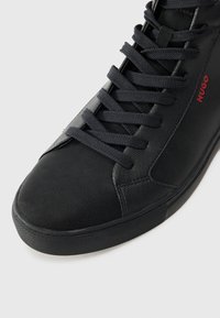 Black high-top sneaker with textured fabric and smooth accents. Features black laces and a red logo, robust rubber sole.
