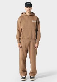 Brun hættejakke med en frontlomme, parret med matchende brune joggingbukser. Har en sort kasket og sneakers med en hvid sål.