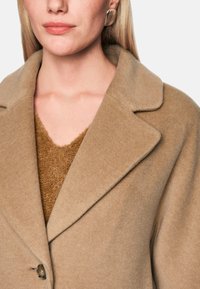Frau, die einen beigen Wollmantel über einem braunen Strickpullover trägt, mit goldenen quadratischen Ohrringen, vor einem einfarbigen Hintergrund.
