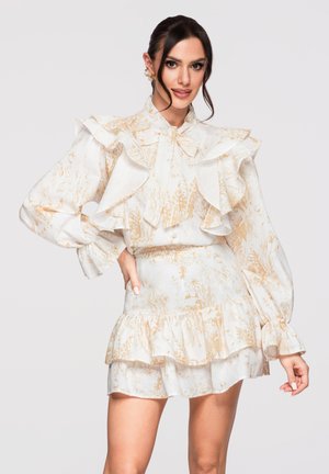 Kobieta pozująca na tle jednolitego tła, ubrana w białą i beżową kwiatową bluzkę z falbankami oraz dopasowaną krótką spódnicę z długimi rękawami i wysokim kołnierzem.