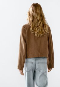 Giacca corta in suede marrone con maniche lunghe e taglio dritto, abbinata a jeans in denim azzurro chiaro, vista da dietro.