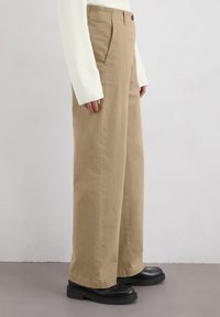 Pantalons larges beige avec une coupe droite, en coton, dotés de poches avant et d'une fermeture à bouton. Associés à des chaussures à plateforme noires.