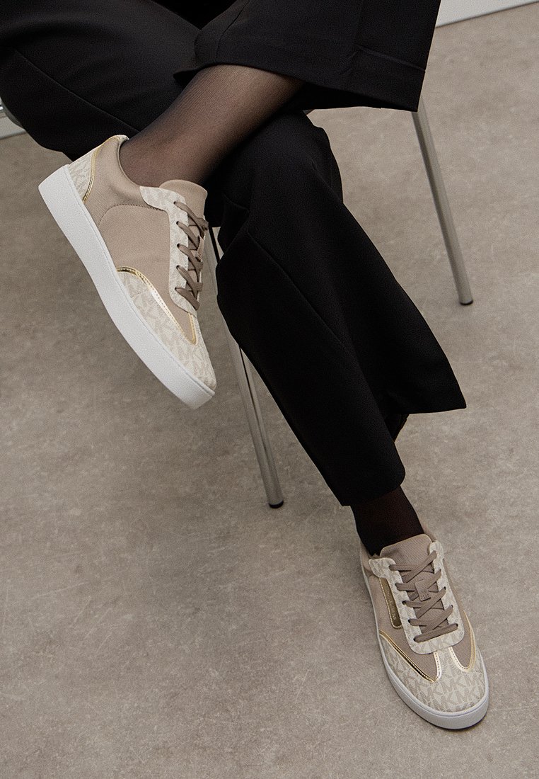 Baskets beiges avec un design texturé, présentant des accents dorés, un laçage à l'avant et une semelle blanche. Portées avec un pantalon noir.