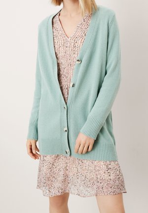 Lys blå cardigan i blød strik med fem knapper, ribbede manchetter og kant. Bæres over en mønstret pink kjole med v-udskæring.