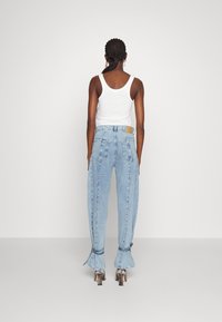 Jeans de mezclilla azul con campana y dobladillo deshilachado, detalles de bolsillo y una etiqueta color beige en la parte posterior. Combinados con una camiseta sin mangas blanca.