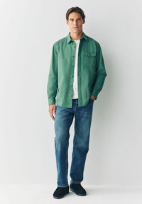 Grüne Button-Down-Hemd, darunter weißes T-Shirt, blaue Jeans und schwarze Schuhe. Das Hemd hat eine einzelne Brusttasche und einen lässigen Schnitt.