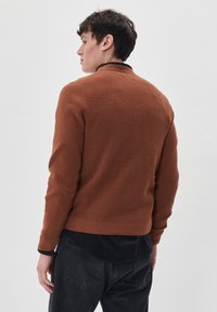 Jersey marrón texturizado con un dobladillo y puños acanalados, que presenta un cuello negro. Llevado sobre una camisa oscura, con un discreto diseño de tejido.