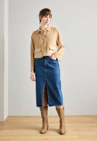 Chemise boutonnée à manches longues de couleur brun clair avec deux poches avant ; jupe en denim de longueur moyenne avec une fente à l'avant ; bottes de style cowboy beiges.