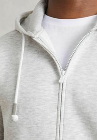 Svijetlo siva zip-up kutija napravljena od mekog materijala, s bijelim patentnim zatvaračem i vezicom. Unutarnji sloj prikazuje bijelu majicu ispod.