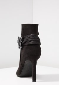 Botín de ante negro con una forma elegante, tacón alto de aguja, correa texturizada envolvente y acento decorativo con joyas en el lateral.