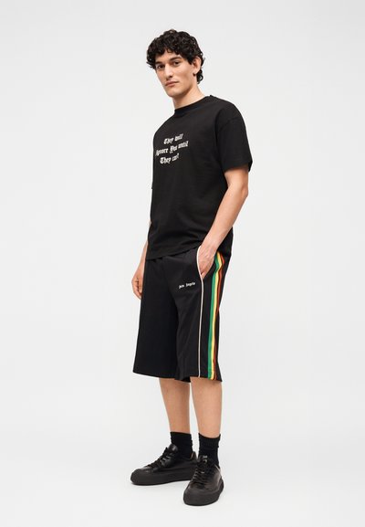 Άνδρας που φοράει μαύρο γραφικό T-shirt και μαύρα σορτς με πράσινες, κίτρινες και κόκκινες ρίγες στο πλάι, στέκεται με το ένα χέρι στην τσέπη μπροστά σε λευκό φόντο.