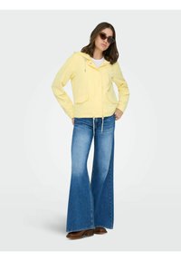 Femme portant une veste à capuche jaune, un jean bleu à jambes larges, des chaussures marron et des lunettes de soleil œil-de-chat, debout les mains dans les poches.