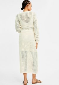 Lang gebreid jurk in crème, met een ruitpatroon, lange mouwen en een getailleerde taille. De stof is lichtgewicht en semi-doorzichtig.