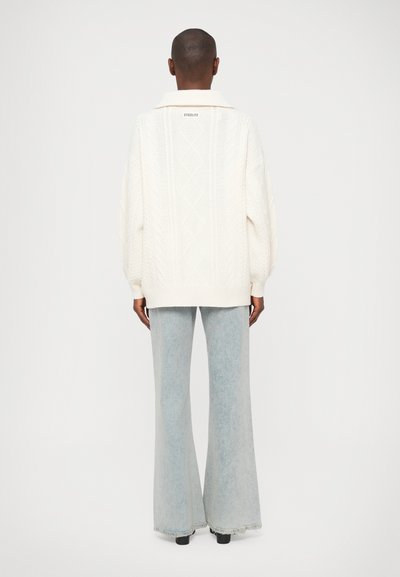 Suéter blanco de punto con un patrón de diamantes y cuello, combinado con jeans acampanados azul claro, mostrado desde la parte de atrás.