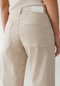 Pantaloni beige con una texture liscia, dotati di due tasche posteriori e un'etichetta con logo bianco in vita. Design classico a cinque tasche.