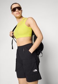 Top deportivo de color amarillo neón combinado con pantalones cortos cargo negros con bolsillos. El modelo lleva una mochila negra y unas gafas de sol oversized.