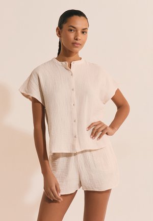 Femme aux cheveux tressés portant un haut beige ample à manches courtes boutonné et un short beige assorti, debout avec une main sur la hanche.