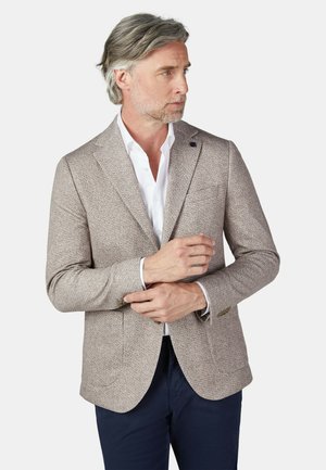 Reifer Mann mit grauem Haar trägt einen beigen strukturierten Blazer, weißes Hemd und marineblaue Hose, richtet seinen Blazerärmel und schaut nach rechts.