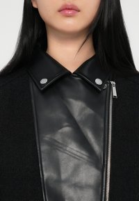 Μαύρο δερμάτινο κολάρο με ασημένιες κουμπώσεις, στρωμένο πάνω σε υφασμάτινη υφή μαύρου χρώματος. Λεπτομέρεια φερμουάρ στη δεξιά πλευρά, κομψό και σύγχρονο σχέδιο.