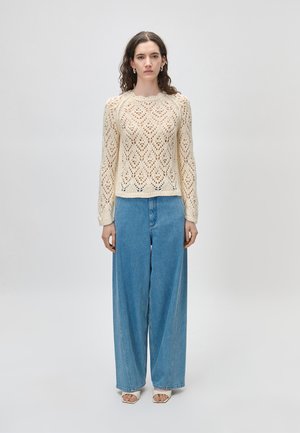 Žena s kudrnatými vlasy, oblečená v krémovém pleteném svetru, širokých modrých džínách a bílých botách se čtvercovou špičkou, stojící před jednoduchým světlým pozadím.