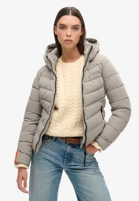 Doudoune grise isolée avec capuche, présentant un design matelassé, une fermeture éclair à l'avant et une accent orange sur les poignets. Portée sur un pull crème en maille torsadée.