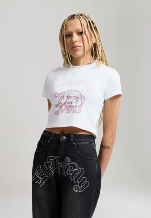 Young person with long blonde braids wearing a white crop top with a panther graphic and black jeans with decorative white text.