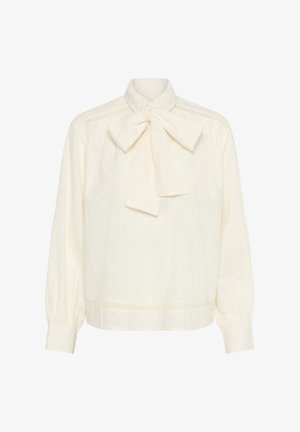Kremowa bluzka z kokardą przy kołnierzu, z teksturowanej tkaniny i koronkowymi detalami wzdłuż ramion i dołu. Długie rękawy.