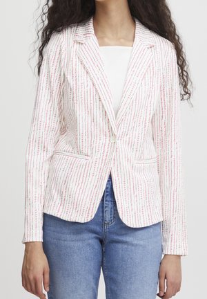 Mujer con una americana blanca con finas rayas verticales rosas sobre una camiseta blanca y vaqueros azules, de pie frente a un fondo liso.