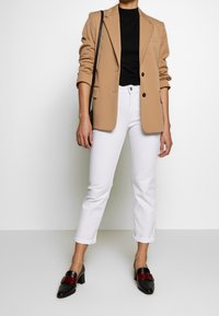 Blazer beige con solapas muesca, top negro ajustado, jeans rectos blancos y mocasines de tacón negros con acentos rojos. Look limpio y bien ajustado.