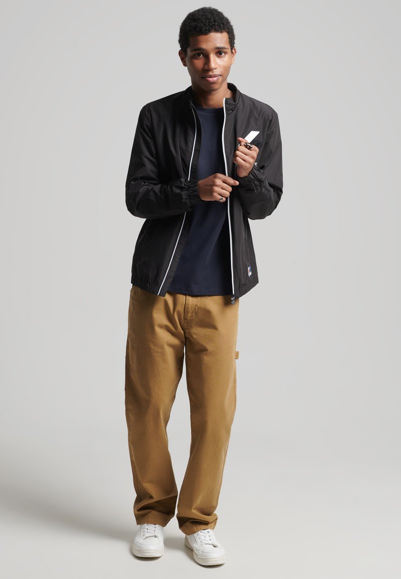 Chaqueta ligera negra con cremallera y puños blancos, combinada con una camisa azul marino y pantalones cargo color beige. El modelo está de pie llevando zapatillas blancas.