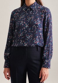 Blusa blu navy con un piccolo motivo floreale bianco, rosso e blu. Caratterizzata da un colletto classico e maniche lunghe a sbuffo. Tessuto di consistenza liscia.