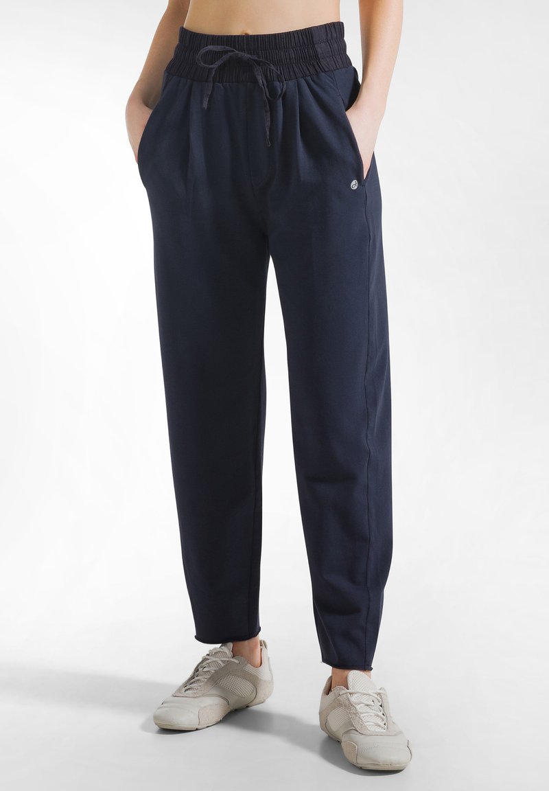 Kobieta ubrana w granatowe spodnie dresowe z elastycznym paskiem w talii i białe trampki, z rękami w kieszeniach, na jednolitym białym tle.