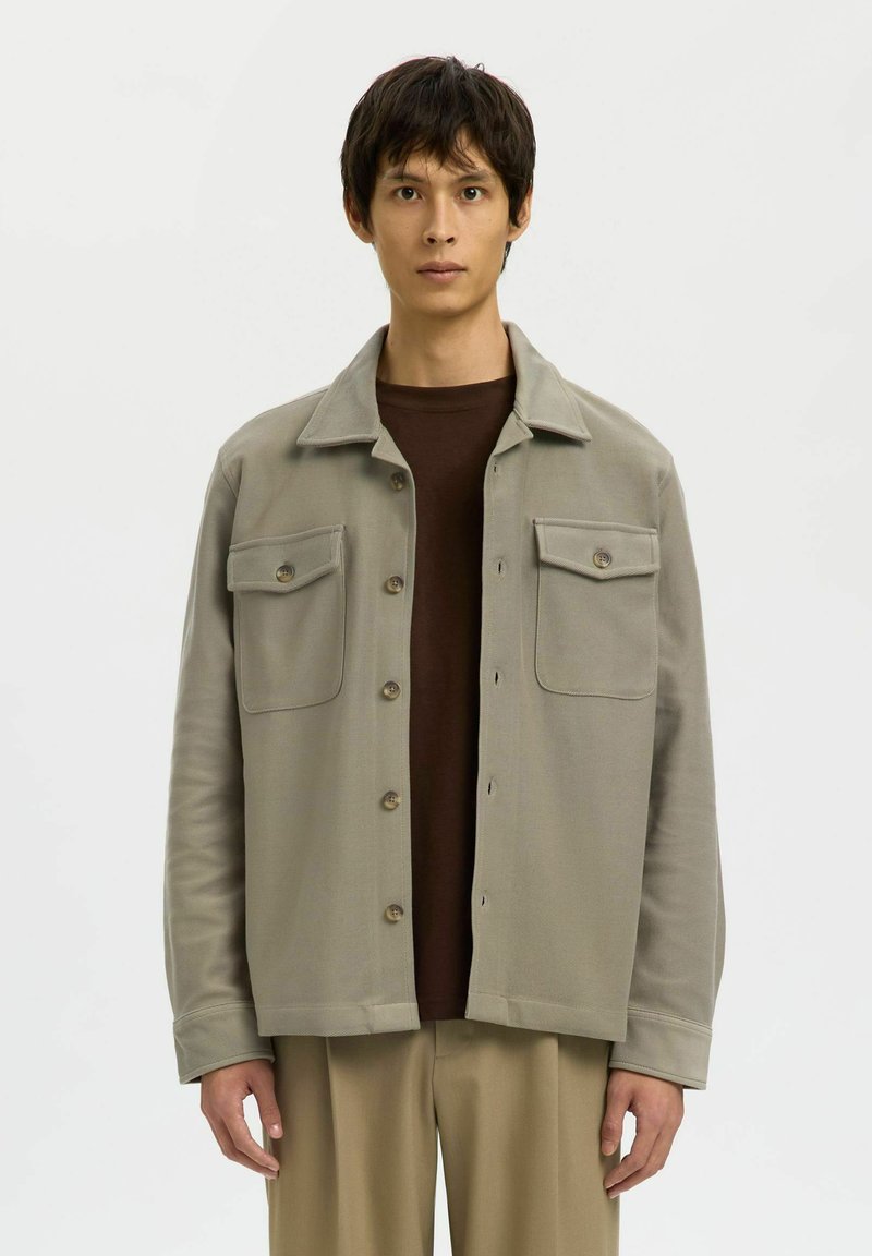 Jonge man in een open beige jas met knoopzakken op de borst, over een bruine shirt en beige broek, staande tegen een effen witte achtergrond.