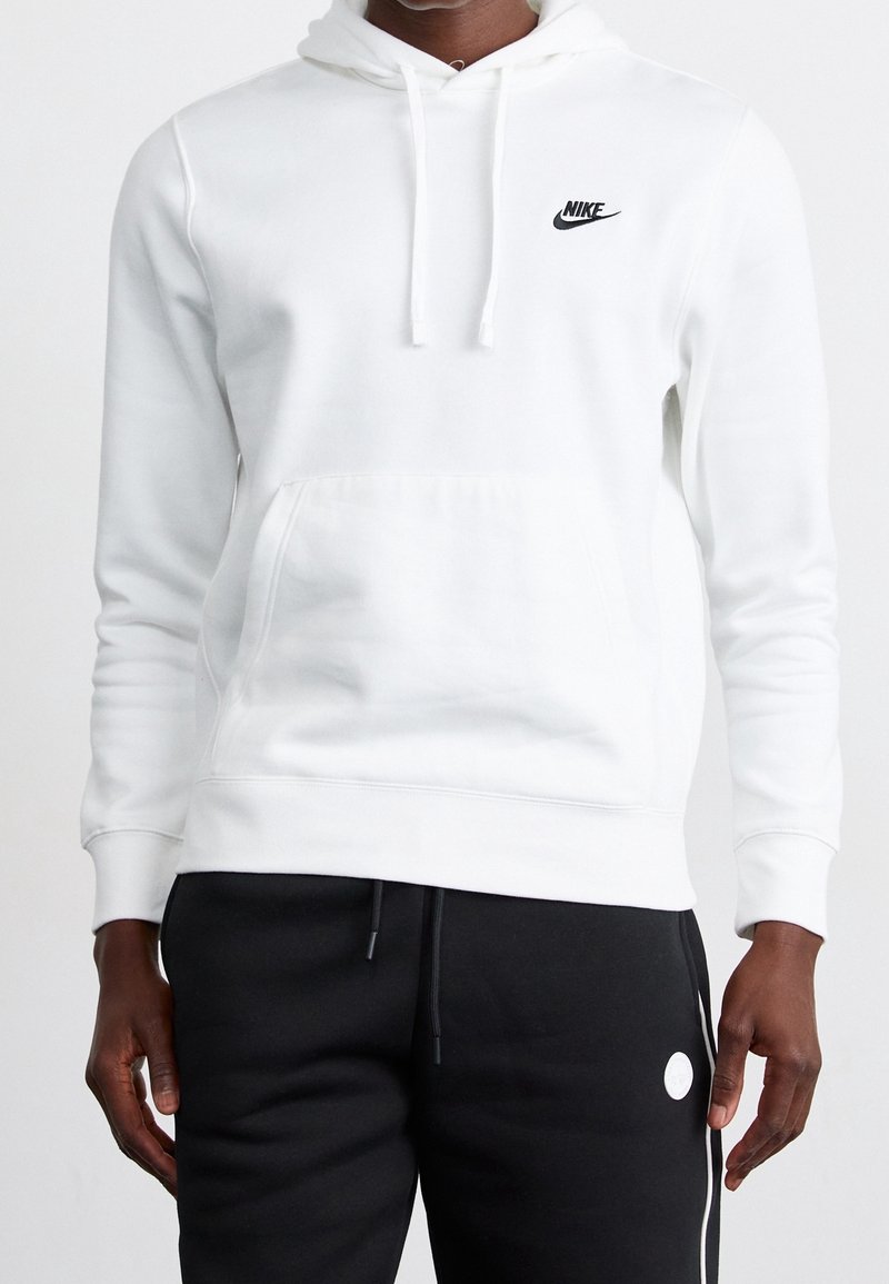 Felpa bianca con cappuccio e tasca frontale, logo Nike nero. Presenta un cappuccio con cordino e polsini a coste. Indossata con pantaloni della tuta neri.