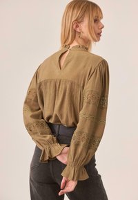 Mujer con una blusa marrón texturizada con inserciones de encaje y puños con volantes, combinada con jeans negros de talle alto, de pie con las manos detrás de la espalda.