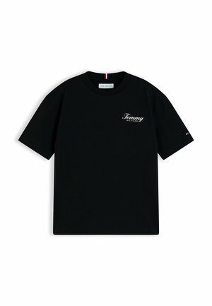 Smilende dreng med krøllet hår iført en sort Tommy Hilfiger t-shirt, blå jeans og beige sneakers, stående med hænderne bag ryggen.
