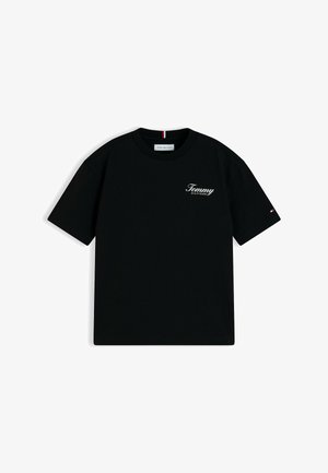 Lächelnder Junge mit lockigem Haar, der ein schwarzes Tommy Hilfiger T-Shirt, blaue Jeans und beige Sneakers trägt, stehend mit verschränkten Händen hinter dem Rücken.