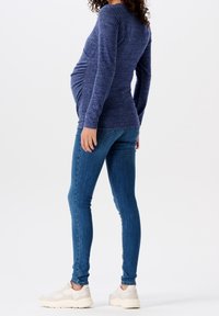 Schwangerenoberteil aus dunkelblauem Strickstoff mit Raffung, kombiniert mit blauen Skinny Jeans und weißen Sneakern. Fokus auf Passform und Komfort.