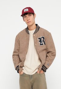 Brun houndstooth bomberjakke med et marineblått "R"-merke og stripet ribbestrikket krage. Bært over en beige T-skjorte og olivengrønne bukser.