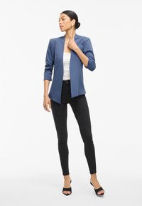 Modrý blejzr s dlouhým rukávem a otevřeným předním designem, doplněný bílým topem a černými skinny džíny. Ať už vypadá skvěle, celkový outfit doladí podpatky.