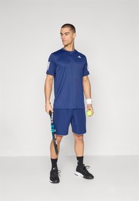 Tumesin sinine sportlik t-särk ja lühikesed püksid, mis on valmistatud kergest kangast. Omab valgeid logo aktsente ja musti tennisekingasid. Hoides tennisereketit ja palli.