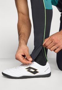 Μαύρο αθλητικό παντελόνι με πράσινες λεπτομέρειες, εφαρμοστό στον αστράγαλο, που ανοίγει με φερμουάρ. Φοράει λευκά αθλητικά παπούτσια με μαύρο λογότυπο.