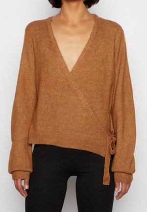 Pull en maille dans un tissu marron texturé et chaud, avec un décolleté en V, des manches longues et un détail à nouer sur le côté. Associé à un pantalon noir.