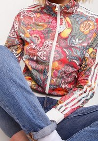 Kolorowa kurtka treningowa Adidas z kwiatowymi wzorami, wysoki kołnierz, zamek błyskawiczny oraz białe paski na ramionach. W zestawie z dżinsami.