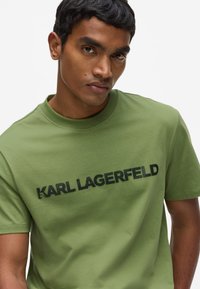 Camiseta de algodón verde con cuello redondo, con el logo negro "KARL LAGERFELD" impreso en el pecho. Corte moderno y textura suave.