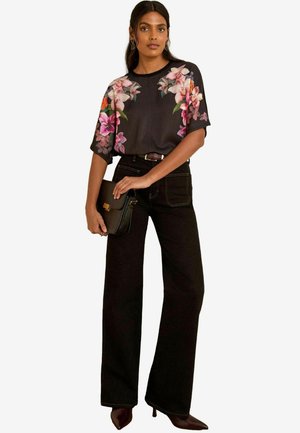 Donna che indossa una blusa nera a maniche corte con stampa floreale, pantaloni neri a vita alta e larghi, tiene in mano una pochette nera e indossa tacchi.