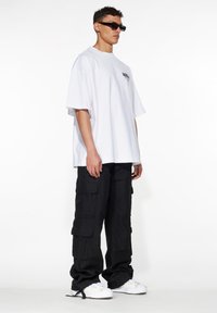 T-shirt bianco oversize, pantaloni cargo neri con tasche e sneakers bianche. Il modello indossa occhiali da sole rettangolari neri. Sfondo semplice.