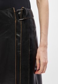 Musta nahkahame, jossa on paksu vyötärönauha hopeisella soljella, kontrastiväriset oranssit tikkauslangat ja kapea siluetti.