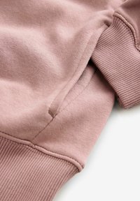 Nahaufnahme eines staubig rosa Sweatshirt-Ärmels mit geripptem Bündchen, sichtbarer Naht und Taschendetail auf weißem Hintergrund.