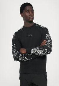 Sweatshirt preto com mangas de padrão floral, punhos e decote canelados. Apresenta um patch de logo no peito. O material parece suave.